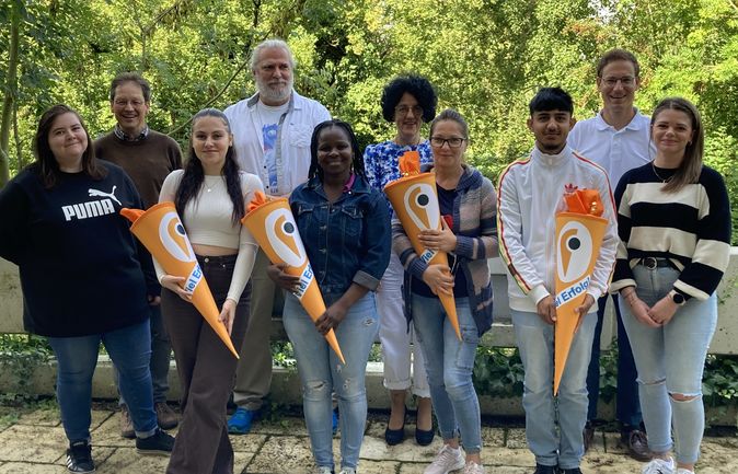 Gruppe aus Mitarbeitenden und neuen Auszubildenden im Freien mit orangen Willkommens-Schultüten