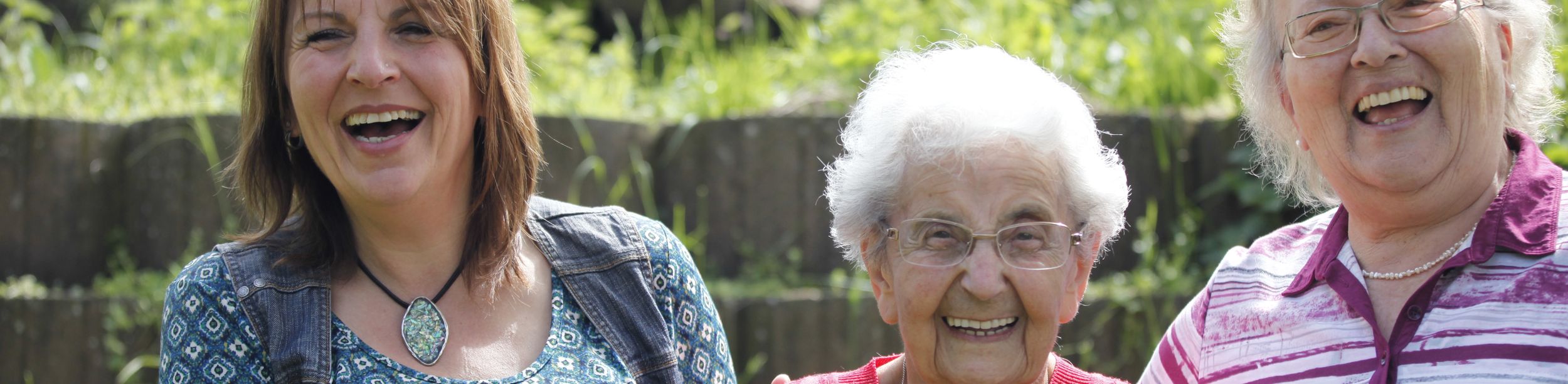 Drei lächelnde Frauen im Garten mit Schafen im Hintergrund